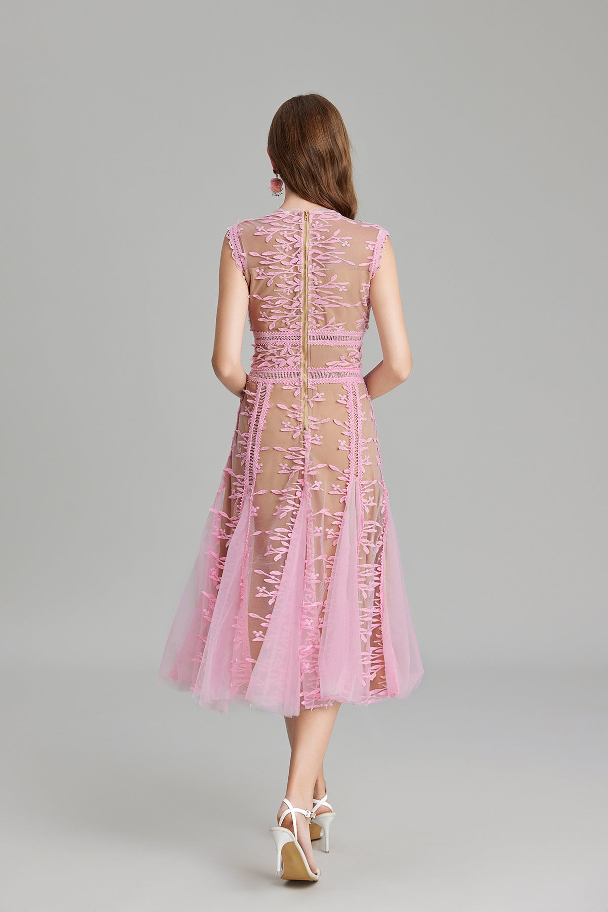 Full-length view of a woman wearing a sleeveless, sheer midi dress in pink and nude, featuring elaborate pink botanical embroidery in vertical panels and a flared tulle skirt.