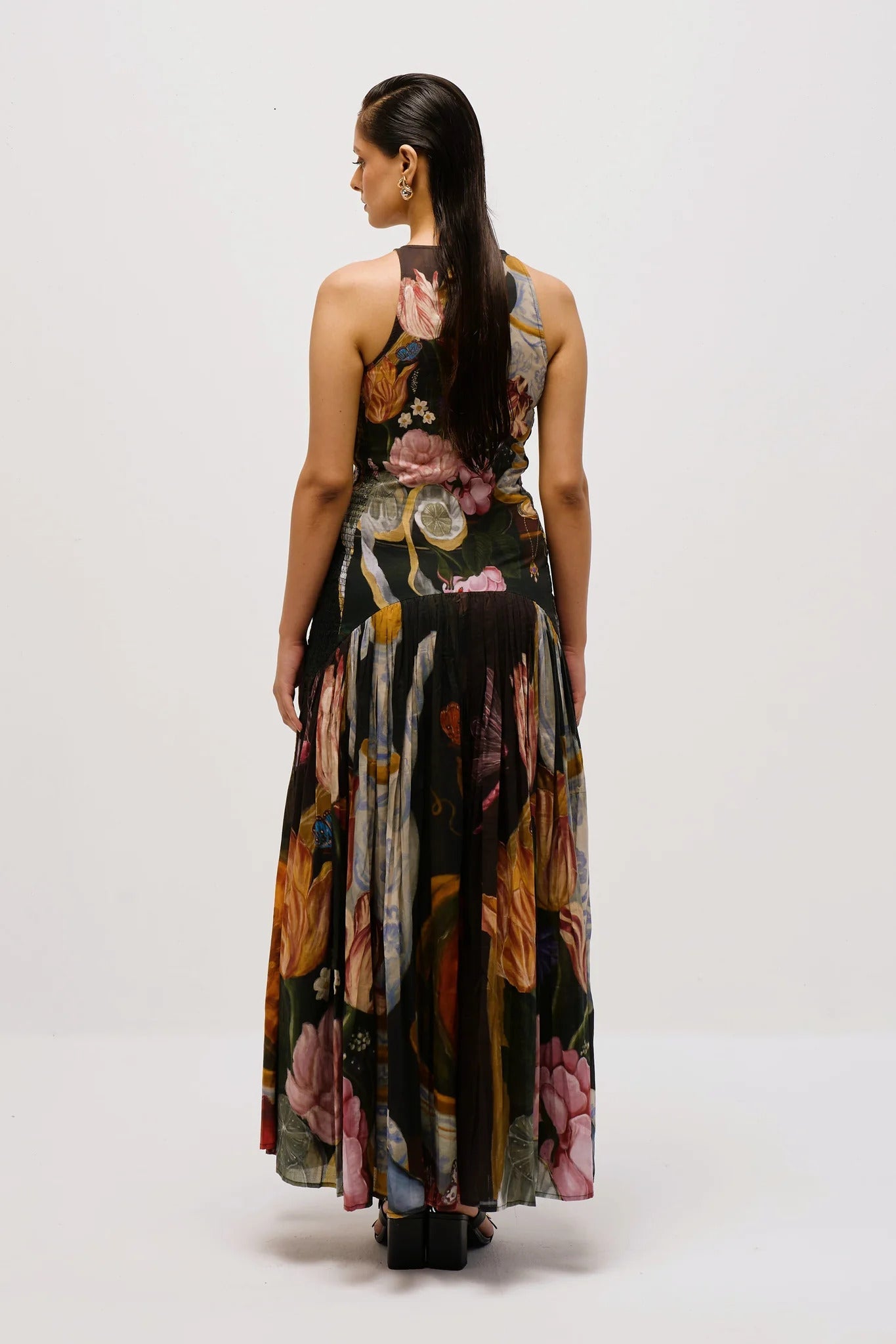 A woman with dark hair slicked back stands against a white background, wearing a full-length, sleeveless halter dress. The dress features a dark, intricate print of flowers, lemons, and antique vases, with a fitted bodice and a long, pleated A-line skirt.