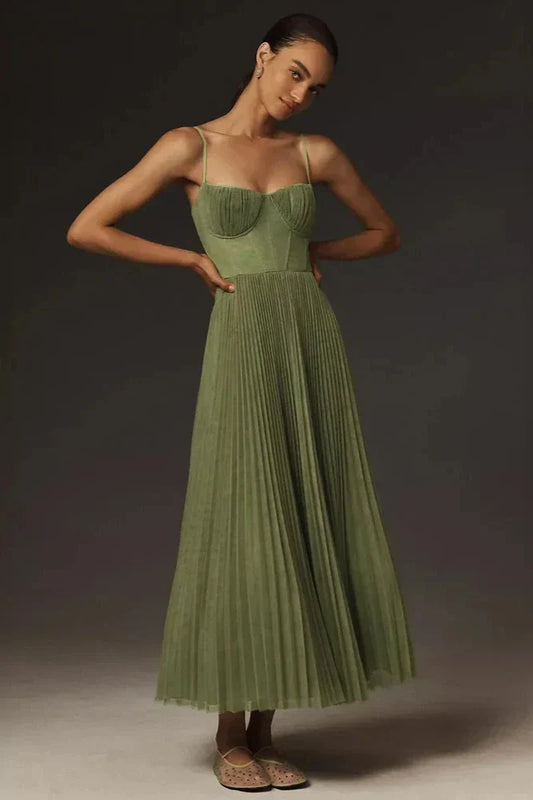 A woman models a sage green midi dress against a dark grey background. The dress has a structured bustier top with delicate spaghetti straps and a full, finely pleated skirt that falls to her lower calf.