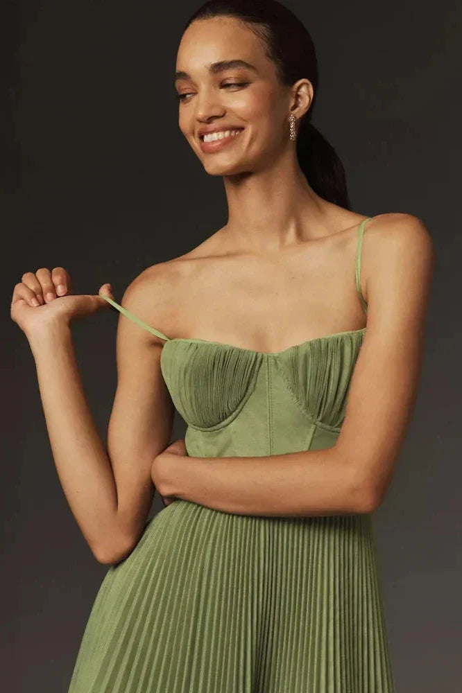 A woman models a sage green midi dress against a dark grey background. The dress has a structured bustier top with delicate spaghetti straps and a full, finely pleated skirt that falls to her lower calf.