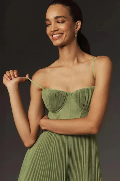 A woman models a sage green midi dress against a dark grey background. The dress has a structured bustier top with delicate spaghetti straps and a full, finely pleated skirt that falls to her lower calf.