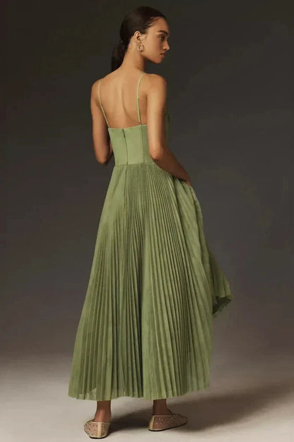 A woman models a sage green midi dress against a dark grey background. The dress has a structured bustier top with delicate spaghetti straps and a full, finely pleated skirt that falls to her lower calf.