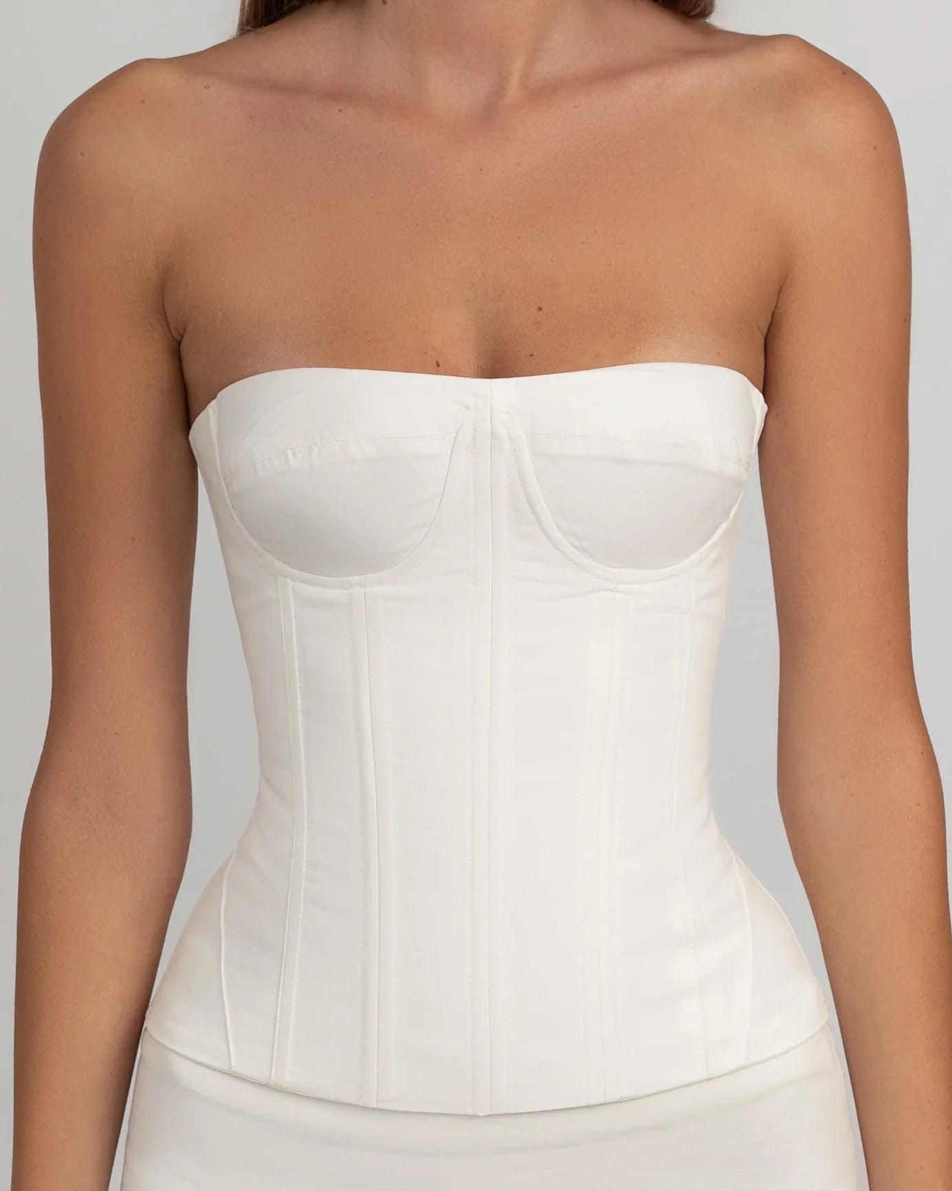 A woman wearing a white, strapless corset-style top with underwire bust cups and visible vertical seaming, paired with a ruffled mini skirt against a light grey background.