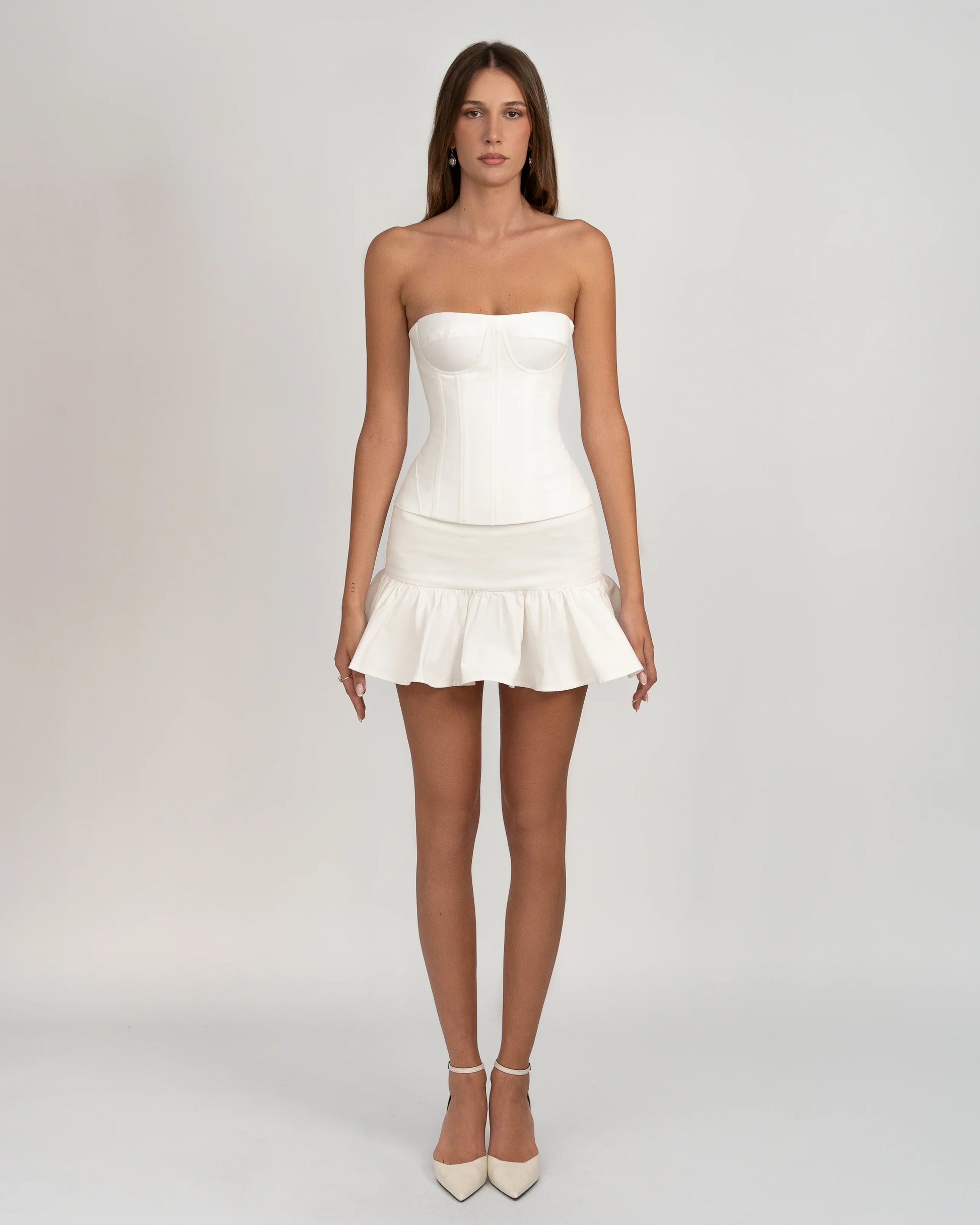 A woman wearing a white, strapless corset-style top with underwire bust cups and visible vertical seaming, paired with a ruffled mini skirt against a light grey background.