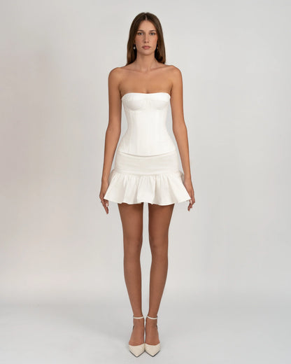 A woman wearing a white, strapless corset-style top with underwire bust cups and visible vertical seaming, paired with a ruffled mini skirt against a light grey background.