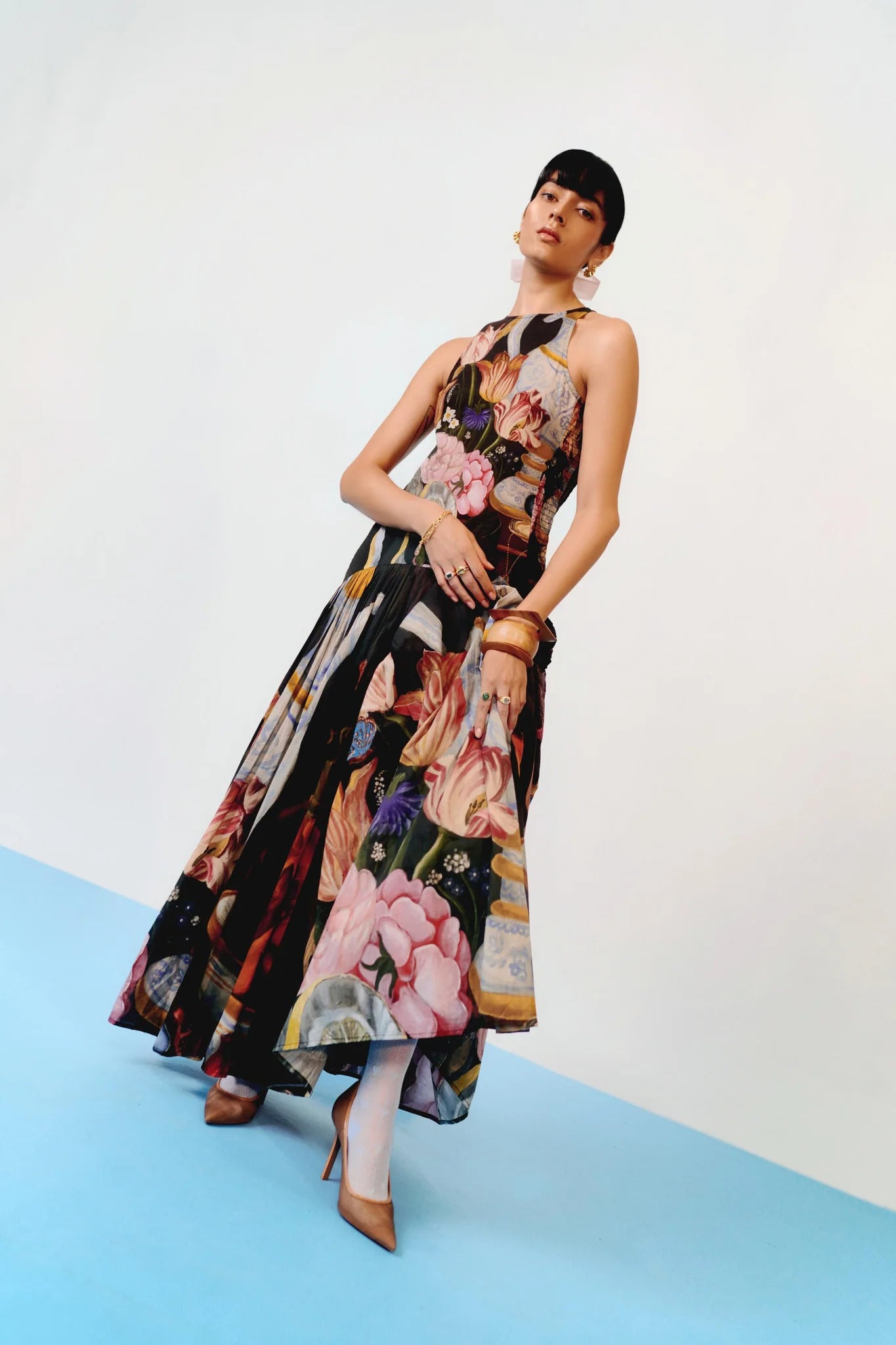A woman with dark hair slicked back stands against a white background, wearing a full-length, sleeveless halter dress. The dress features a dark, intricate print of flowers, lemons, and antique vases, with a fitted bodice and a long, pleated A-line skirt.