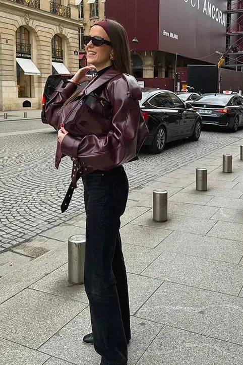 A woman wearing a burgundy oversized faux leather jacket with wide lapels, belted cuffs, and voluminous sleeves, styled over a black turtleneck in a city setting.
