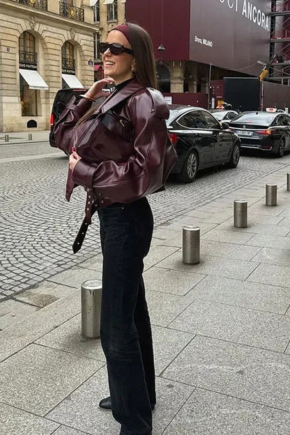 A woman wearing a burgundy oversized faux leather jacket with wide lapels, belted cuffs, and voluminous sleeves, styled over a black turtleneck in a city setting.