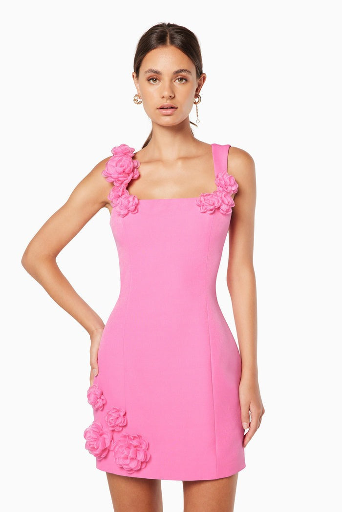 A woman models a white sleeveless A-line mini dress with a square neckline against a white background. The dress is detailed with white 3D floral appliqués on the straps and the left hip.