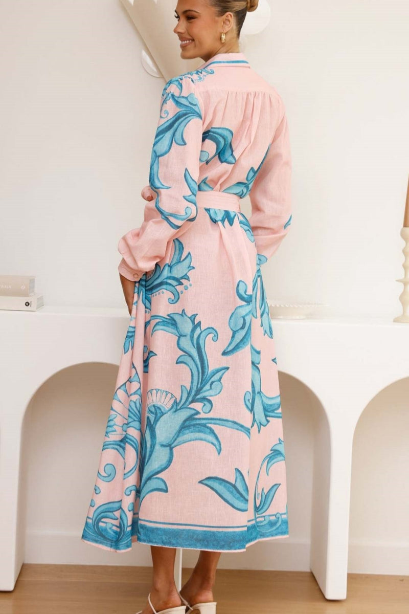 A woman models a long-sleeve maxi shirtdress in pink with a large-scale blue baroque scroll print. The dress features a collar, a full button-front placket, bishop sleeves, and a matching tie-waist belt. She is standing in a minimalist white room.
