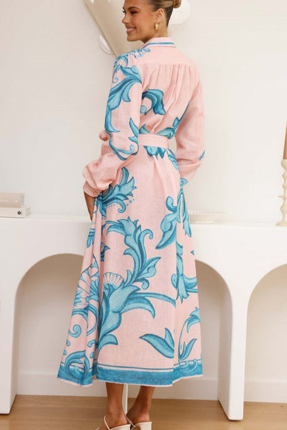 A woman models a long-sleeve maxi shirtdress in pink with a large-scale blue baroque scroll print. The dress features a collar, a full button-front placket, bishop sleeves, and a matching tie-waist belt. She is standing in a minimalist white room.