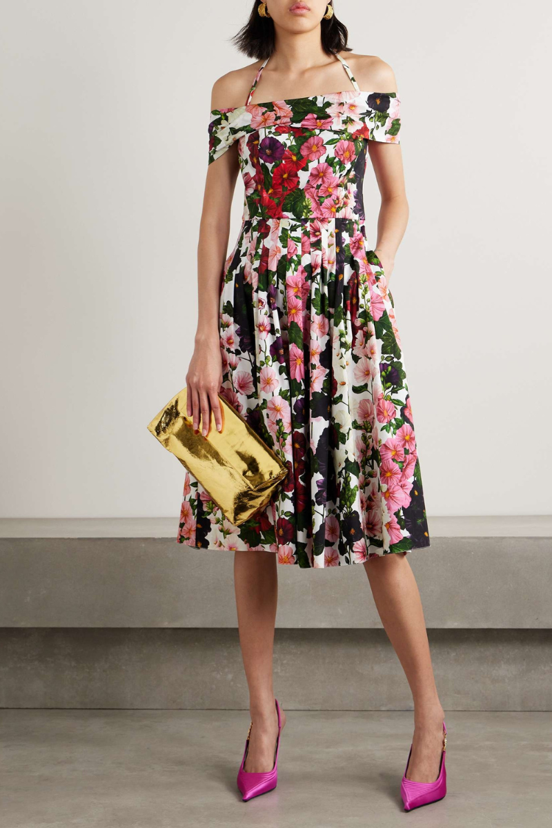 A woman models a knee-length, off-the-shoulder dress with a halter strap, featuring a vibrant floral print on a white background. She holds a gold clutch and wears pink satin heels.