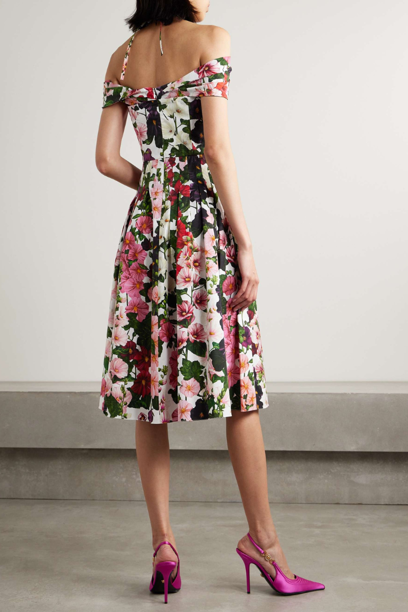 A woman models a knee-length, off-the-shoulder dress with a halter strap, featuring a vibrant floral print on a white background. She holds a gold clutch and wears pink satin heels.