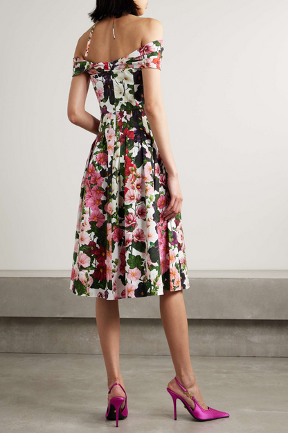 A woman models a knee-length, off-the-shoulder dress with a halter strap, featuring a vibrant floral print on a white background. She holds a gold clutch and wears pink satin heels.