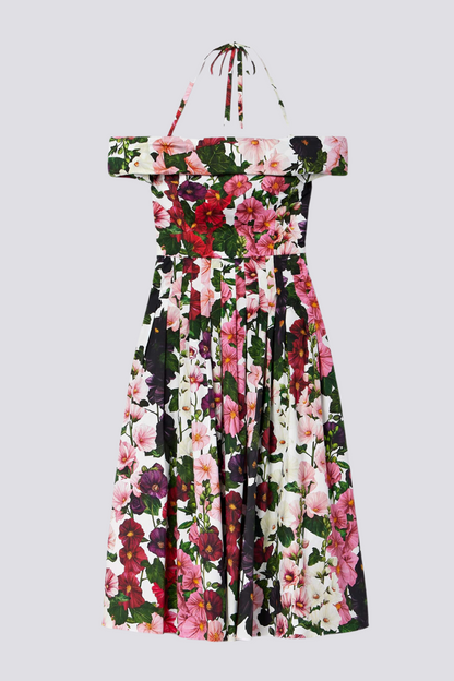 A woman models a knee-length, off-the-shoulder dress with a halter strap, featuring a vibrant floral print on a white background. She holds a gold clutch and wears pink satin heels.