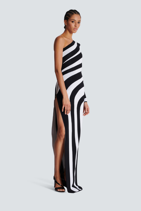 A model stands in profile wearing a form-fitting, one-shoulder maxi dress with a single long sleeve, featuring a bold black and white diagonal stripe pattern and a high thigh slit.
