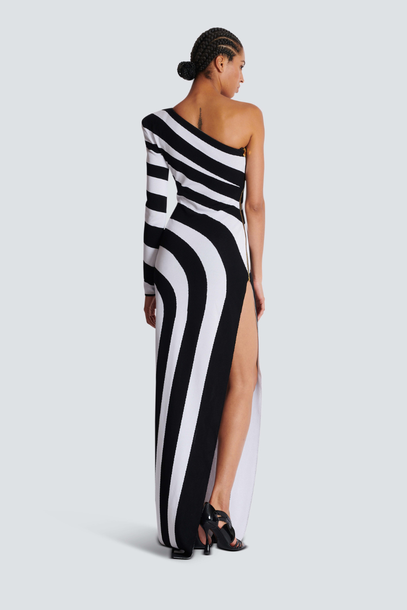 A model stands in profile wearing a form-fitting, one-shoulder maxi dress with a single long sleeve, featuring a bold black and white diagonal stripe pattern and a high thigh slit.