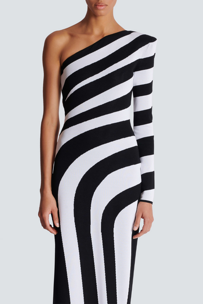 Black - A model stands in profile wearing a form-fitting, one-shoulder maxi dress with a single long sleeve, featuring a bold black and white diagonal stripe pattern and a high thigh slit.