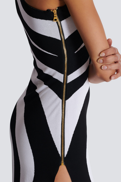 Black - A model stands in profile wearing a form-fitting, one-shoulder maxi dress with a single long sleeve, featuring a bold black and white diagonal stripe pattern and a high thigh slit.