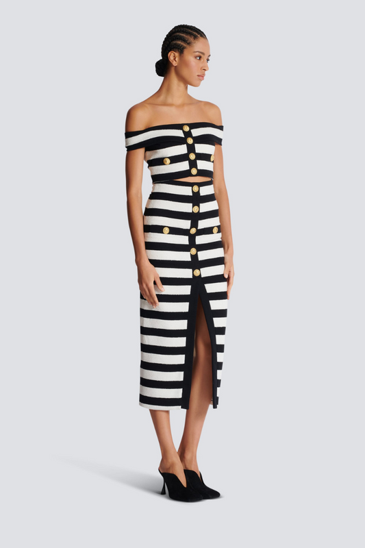 Black - Full-length side view of a model wearing a black and white striped two-piece dress, featuring an off-the-shoulder crop top and a high-waisted midi skirt with a front slit and gold buttons.