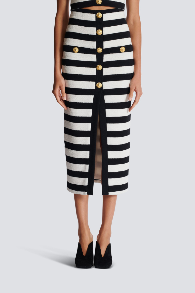 Black - Full-length side view of a model wearing a black and white striped two-piece dress, featuring an off-the-shoulder crop top and a high-waisted midi skirt with a front slit and gold buttons.