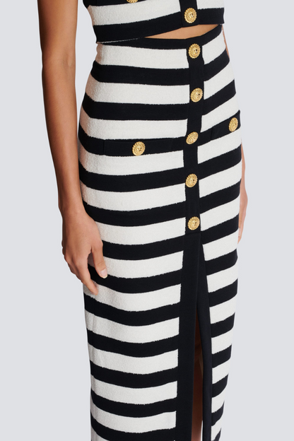 Black - Full-length side view of a model wearing a black and white striped two-piece dress, featuring an off-the-shoulder crop top and a high-waisted midi skirt with a front slit and gold buttons.