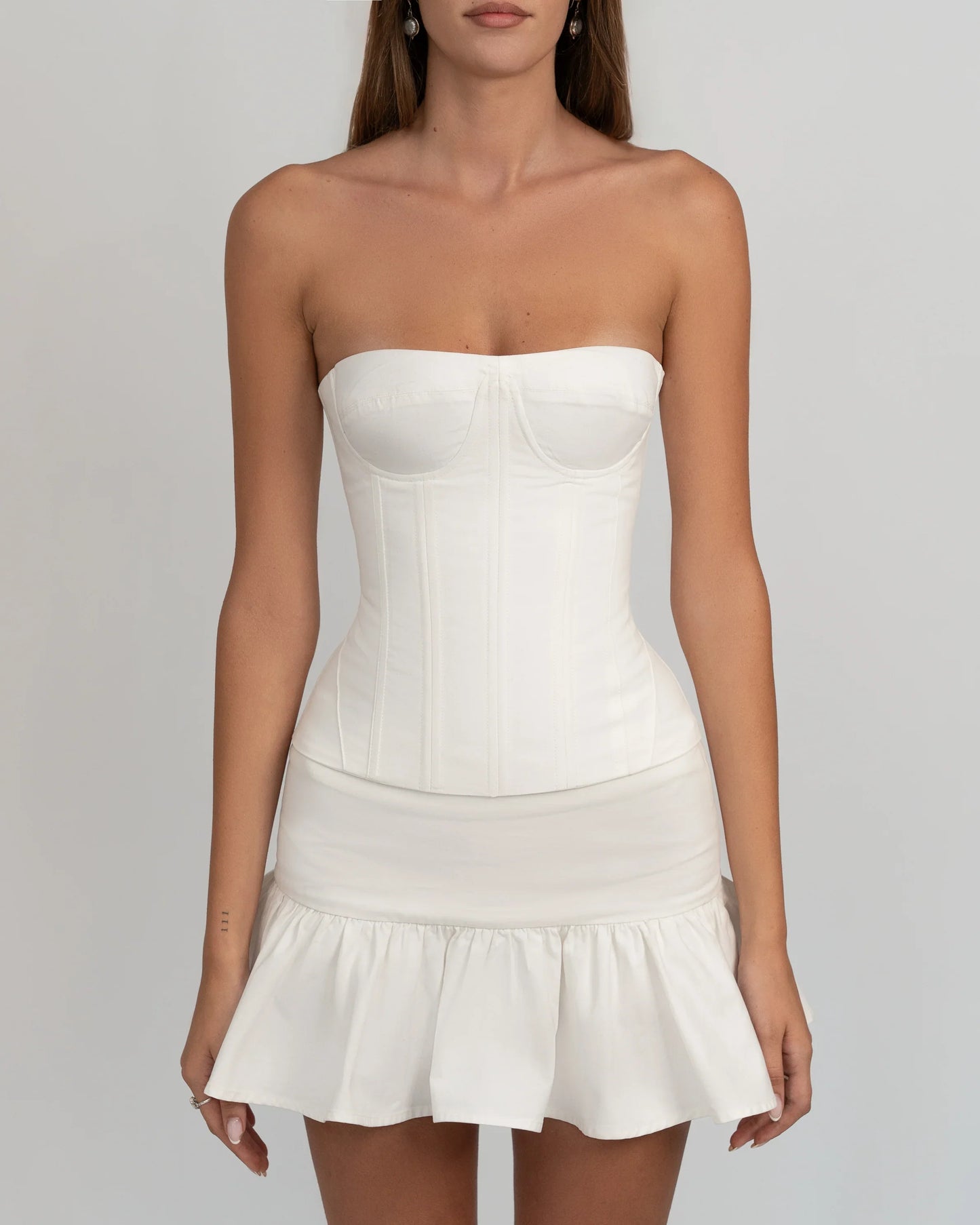 A woman wearing a white, strapless corset-style top with underwire bust cups and visible vertical seaming, paired with a ruffled mini skirt against a light grey background.