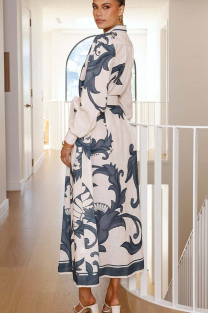 A woman models a long-sleeve maxi shirtdress in pink with a large-scale blue baroque scroll print. The dress features a collar, a full button-front placket, bishop sleeves, and a matching tie-waist belt. She is standing in a minimalist white room.