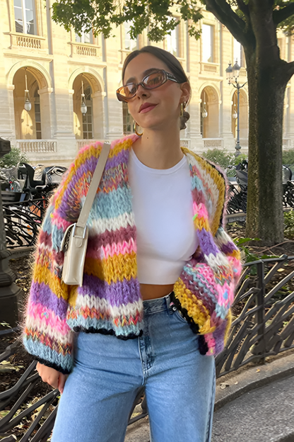Rainbow - Woman wearing a vibrant, multicoloured striped chunky knit cropped cardigan layered over a white top, standing outdoors.
