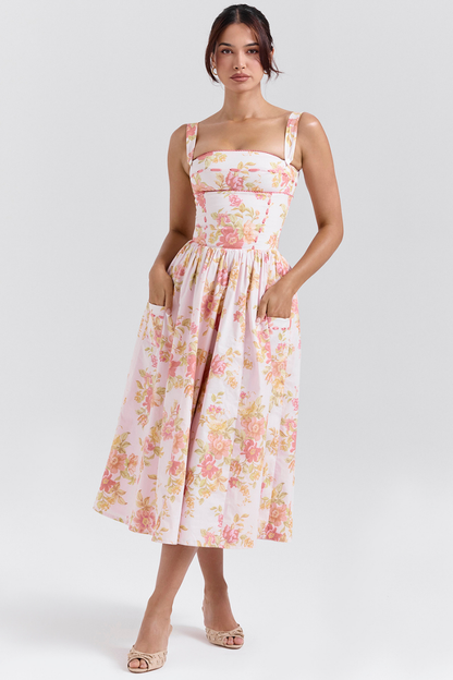 A woman stands against a light gray background wearing a floral print midi dress with a structured corset bodice, wide shoulder straps, and a full A-line skirt with her hands in the side pockets.