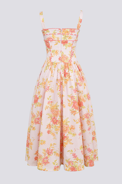 A woman stands against a light gray background wearing a floral print midi dress with a structured corset bodice, wide shoulder straps, and a full A-line skirt with her hands in the side pockets.
