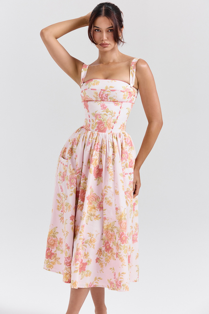 A woman stands against a light gray background wearing a floral print midi dress with a structured corset bodice, wide shoulder straps, and a full A-line skirt with her hands in the side pockets.
