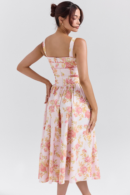 A woman stands against a light gray background wearing a floral print midi dress with a structured corset bodice, wide shoulder straps, and a full A-line skirt with her hands in the side pockets.
