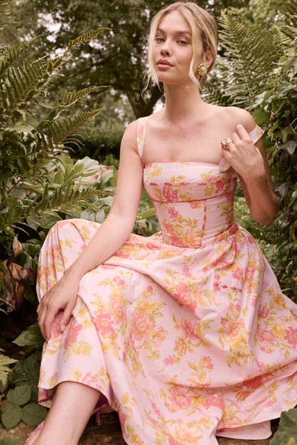A woman stands against a light gray background wearing a floral print midi dress with a structured corset bodice, wide shoulder straps, and a full A-line skirt with her hands in the side pockets.