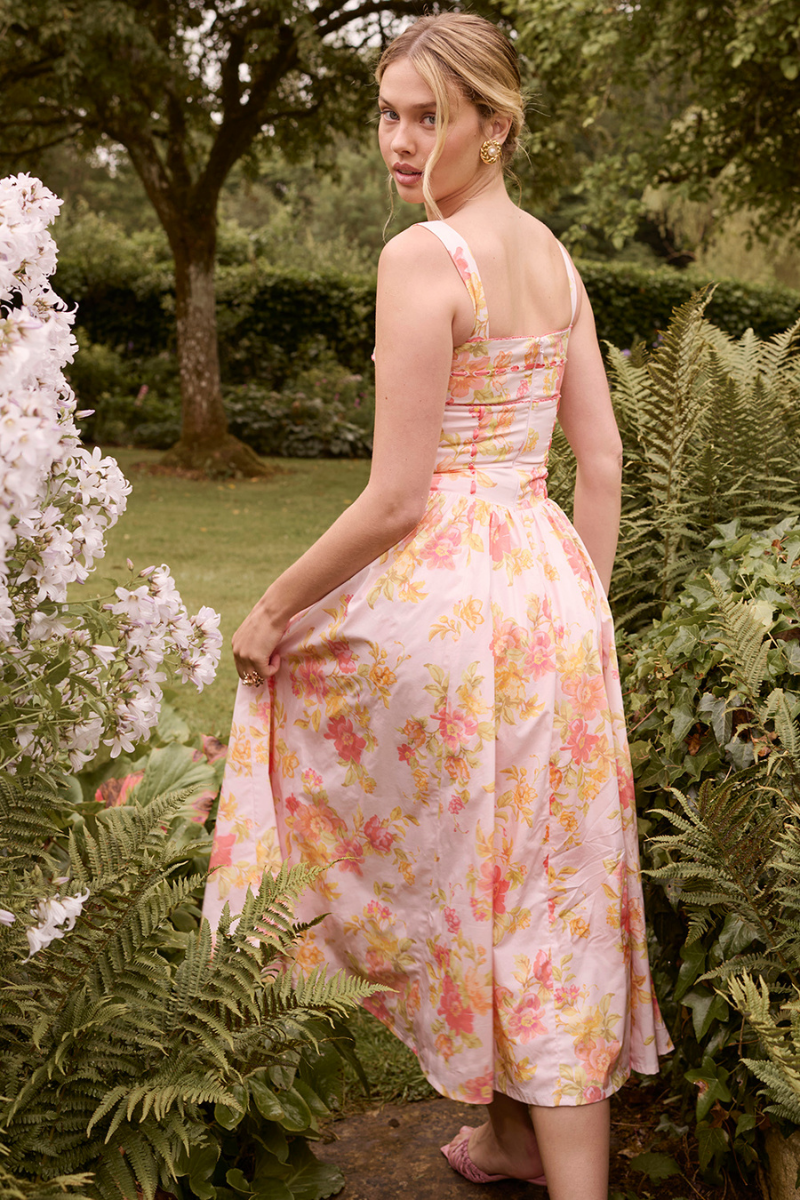 A woman stands against a light gray background wearing a floral print midi dress with a structured corset bodice, wide shoulder straps, and a full A-line skirt with her hands in the side pockets.