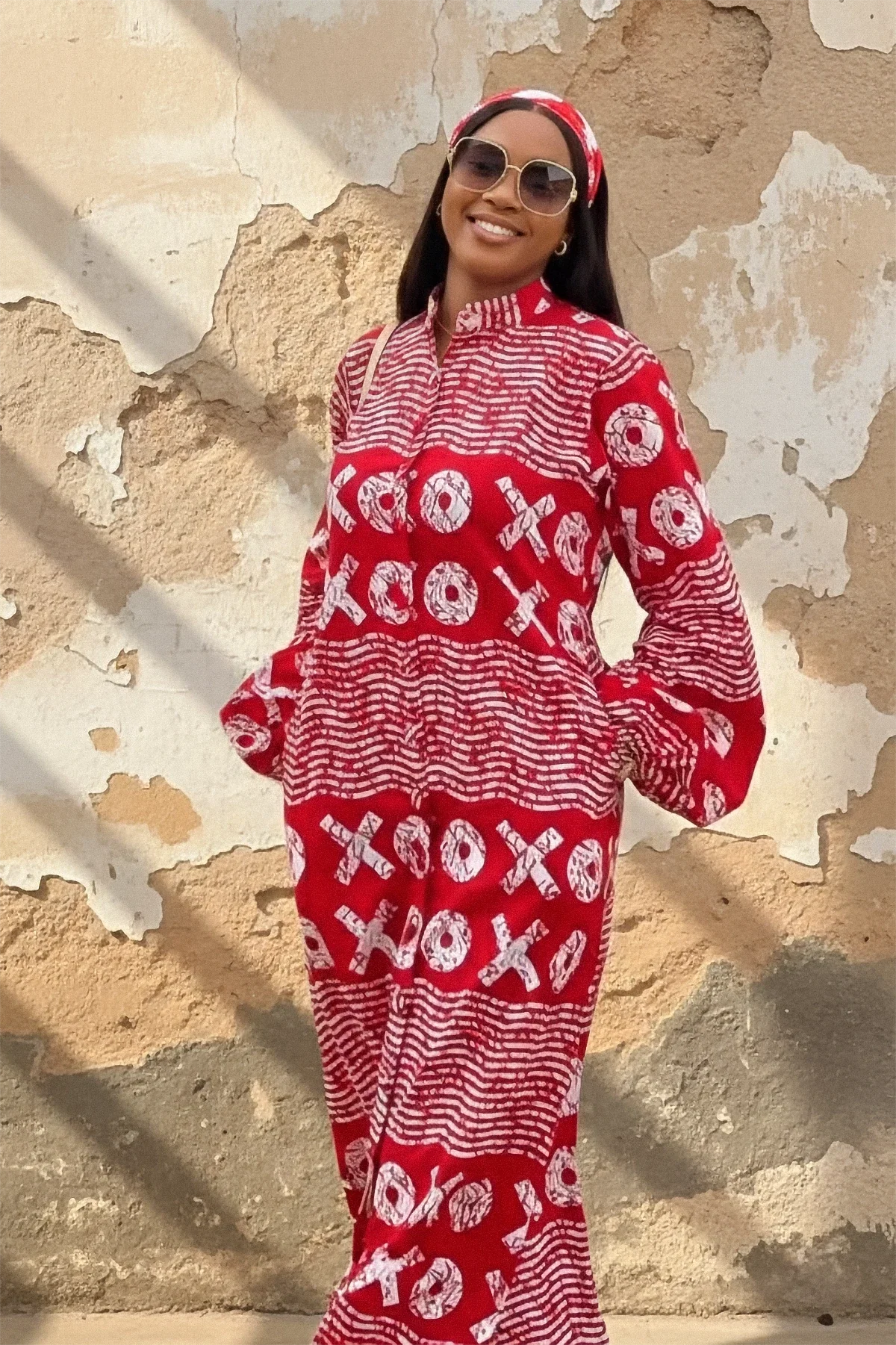 A woman wearing a red and white XOXO print maxi dress with a high neck, long flared sleeves, and a matching headband. She is smiling and standing with her arms out against a weathered beige wall.