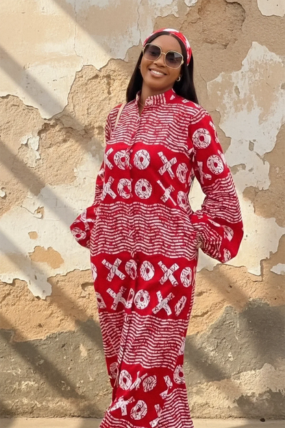 A woman wearing a red and white XOXO print maxi dress with a high neck, long flared sleeves, and a matching headband. She is smiling and standing with her arms out against a weathered beige wall.