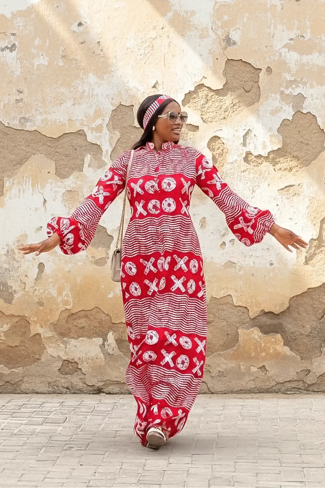 A woman wearing a red and white XOXO print maxi dress with a high neck, long flared sleeves, and a matching headband. She is smiling and standing with her arms out against a weathered beige wall.
