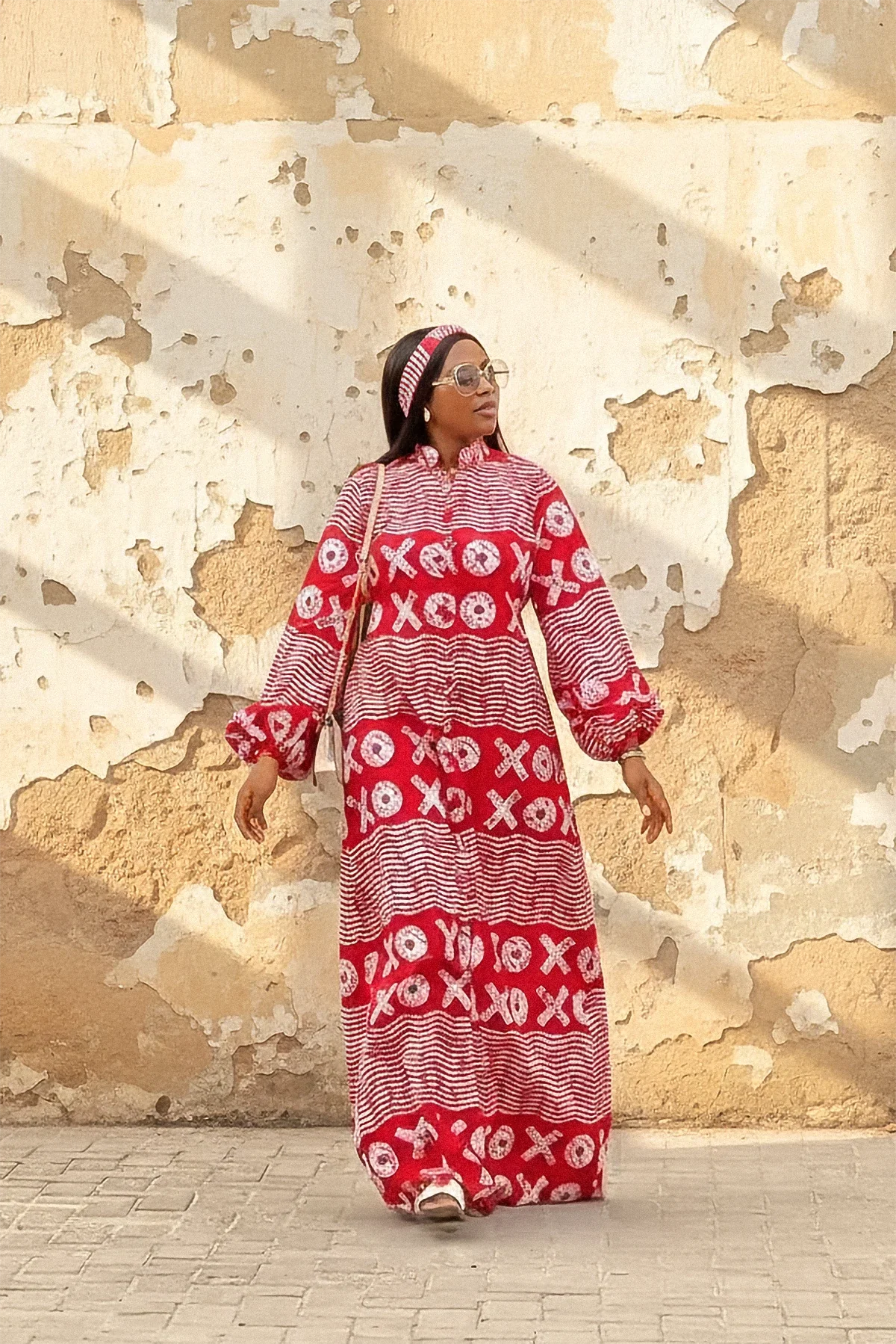 A woman wearing a red and white XOXO print maxi dress with a high neck, long flared sleeves, and a matching headband. She is smiling and standing with her arms out against a weathered beige wall.