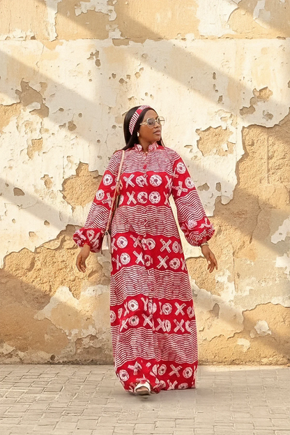 A woman wearing a red and white XOXO print maxi dress with a high neck, long flared sleeves, and a matching headband. She is smiling and standing with her arms out against a weathered beige wall.