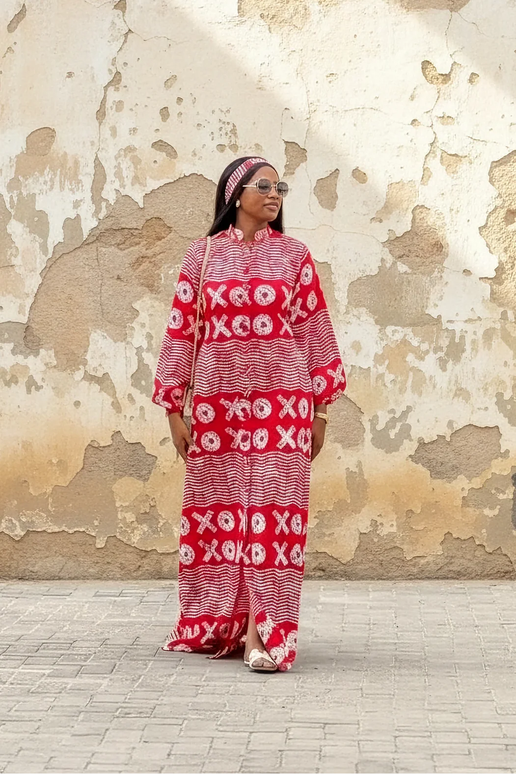 A woman wearing a red and white XOXO print maxi dress with a high neck, long flared sleeves, and a matching headband. She is smiling and standing with her arms out against a weathered beige wall.