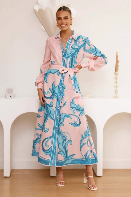 A woman models a long-sleeve maxi shirtdress in pink with a large-scale blue baroque scroll print. The dress features a collar, a full button-front placket, bishop sleeves, and a matching tie-waist belt. She is standing in a minimalist white room.