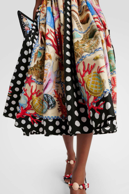 Full-length view of a woman wearing a mixed-print midi dress with a scenic-print square-neck bodice, polka dot straps, and a full A-line skirt featuring seashell and polka dot panels.