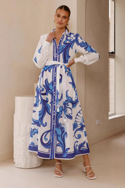 A woman models a long-sleeve maxi shirtdress in pink with a large-scale blue baroque scroll print. The dress features a collar, a full button-front placket, bishop sleeves, and a matching tie-waist belt. She is standing in a minimalist white room.