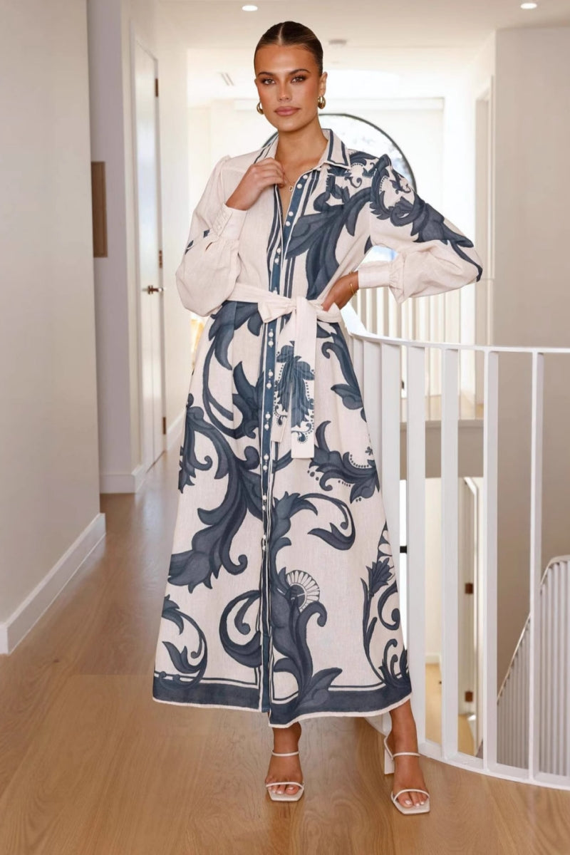 A woman models a long-sleeve maxi shirtdress in pink with a large-scale blue baroque scroll print. The dress features a collar, a full button-front placket, bishop sleeves, and a matching tie-waist belt. She is standing in a minimalist white room.