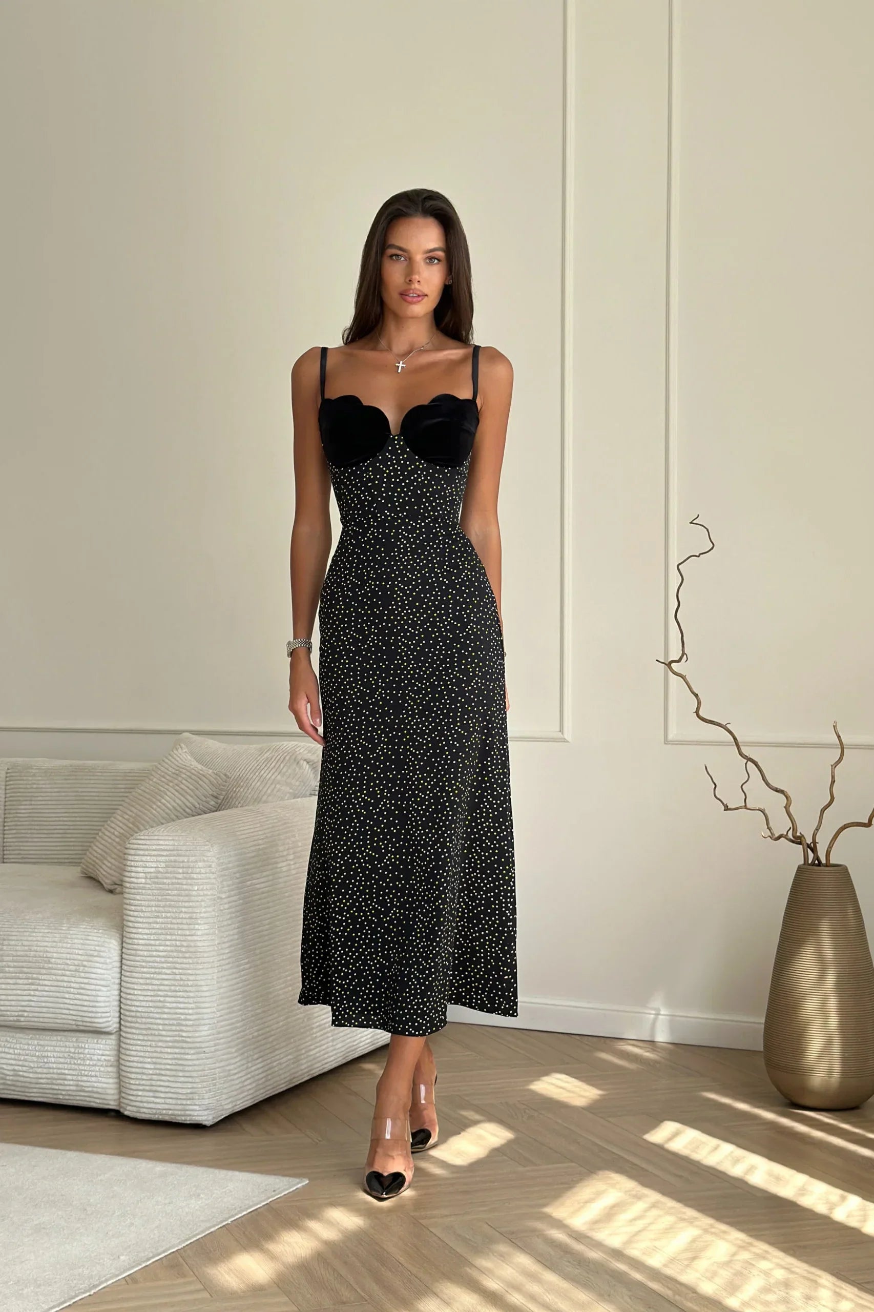 A woman with long brown hair leans against a white wall, wearing a black midi slip dress with white polka dots. The dress features thin spaghetti straps and a contrasting black scalloped bustier top.