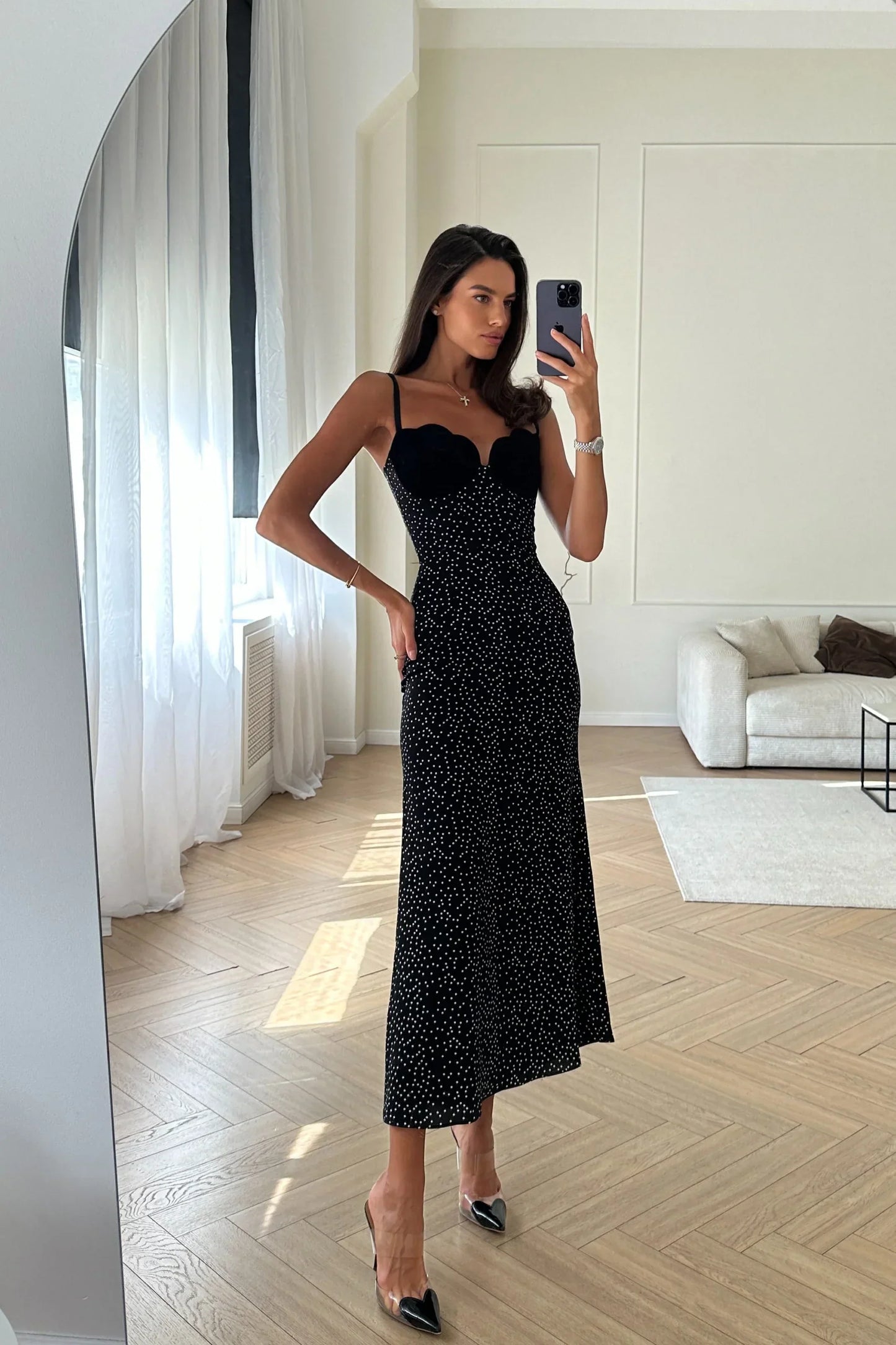 A woman with long brown hair leans against a white wall, wearing a black midi slip dress with white polka dots. The dress features thin spaghetti straps and a contrasting black scalloped bustier top.