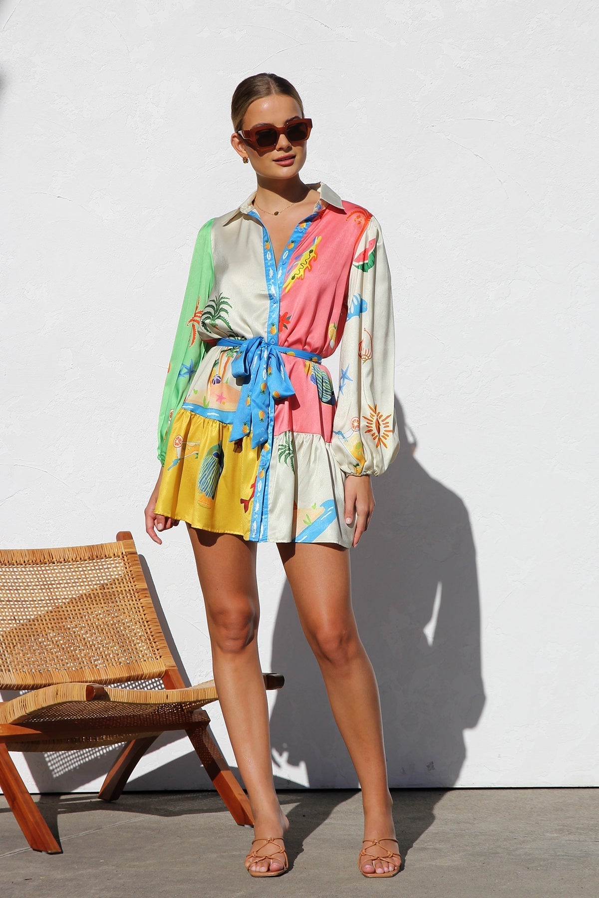 A woman with her hair in a sleek bun smiles while wearing a multi-patterned, long-sleeve tropical print mini shirt dress, standing against a white textured wall.