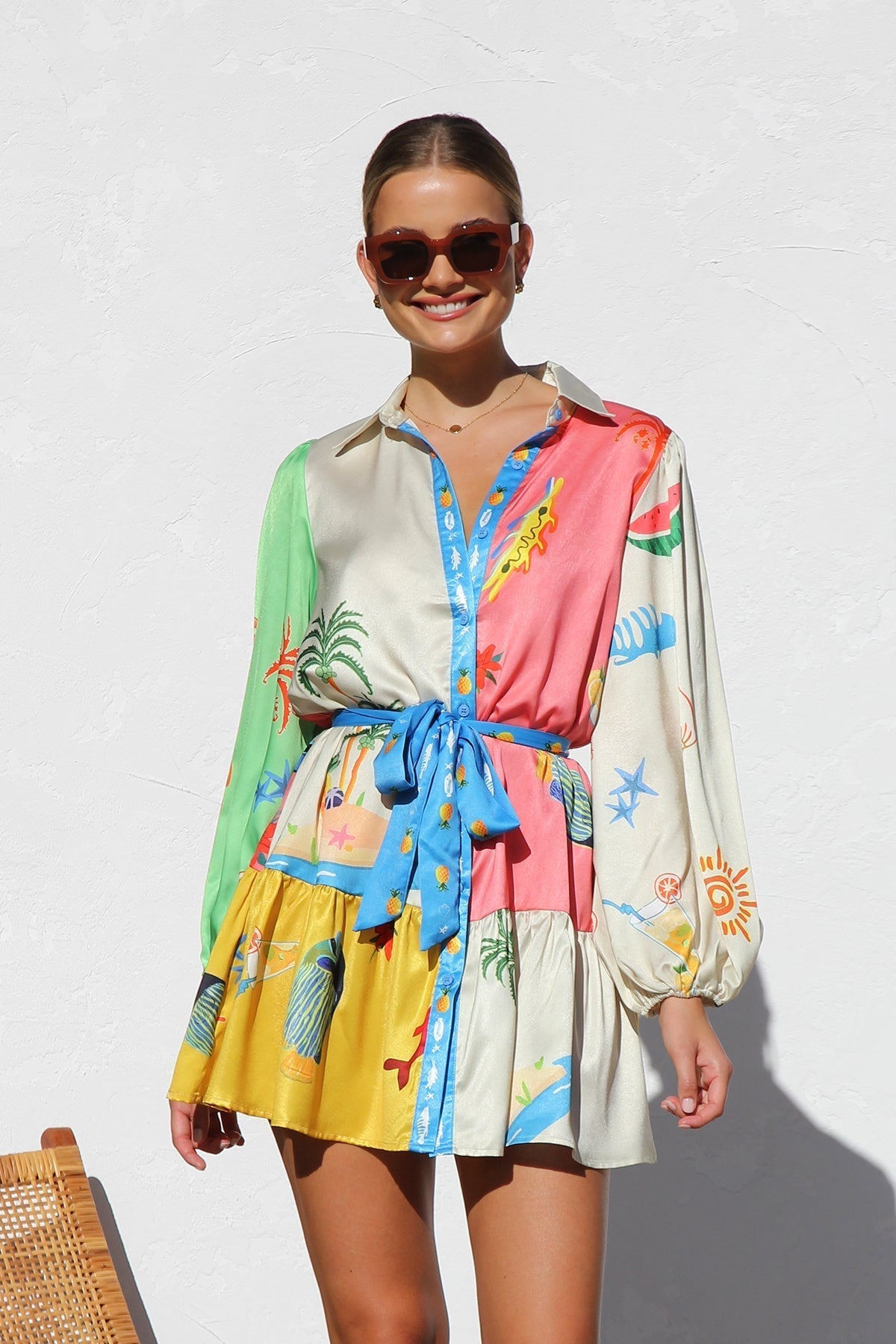 A woman with blonde hair in a bun smiles while wearing a colorful, long-sleeve mini shirt dress with a tropical print, belted at the waist, standing against a white textured wall.