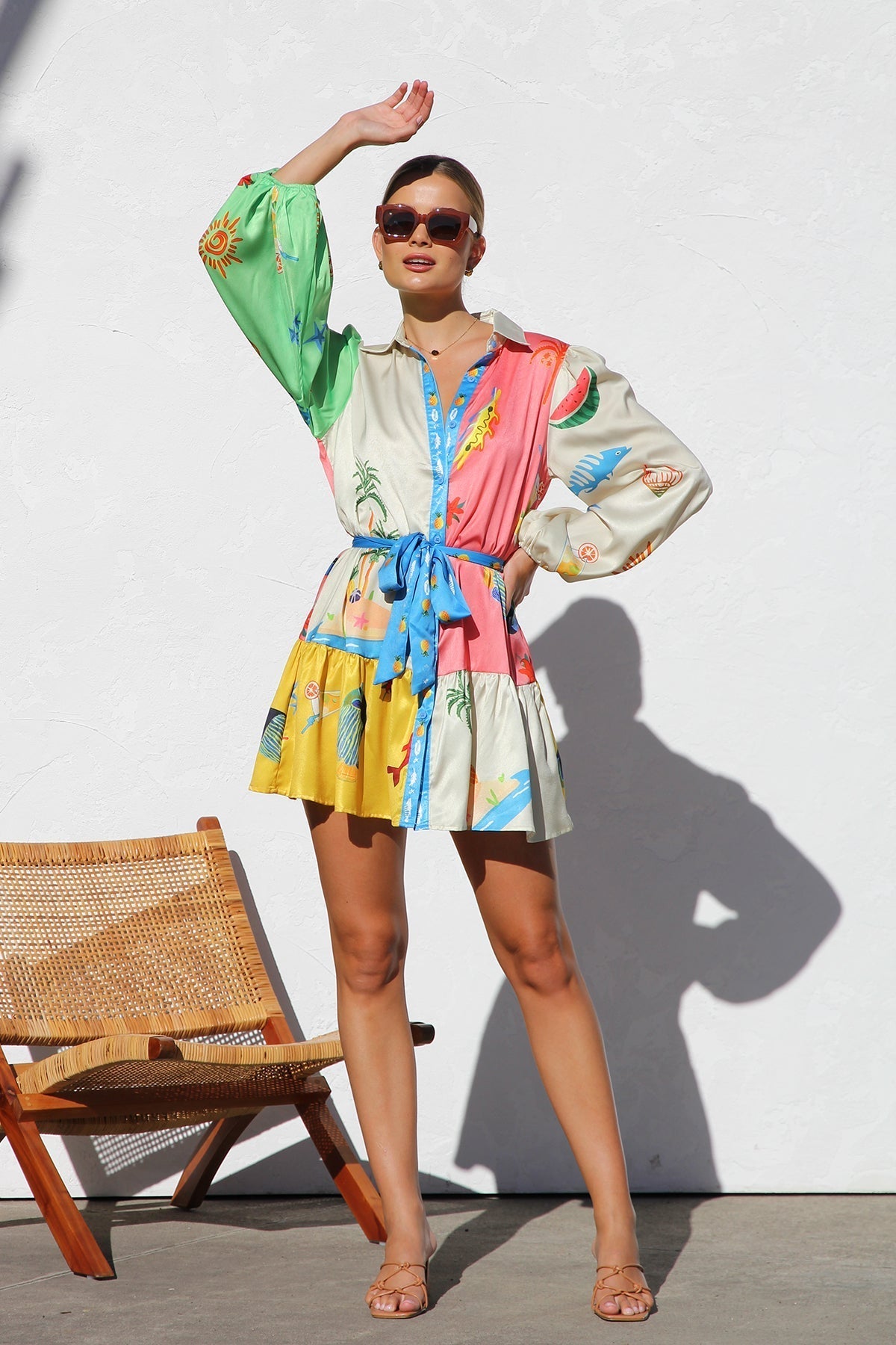 A woman with blonde hair in a bun smiles while wearing a colorful, long-sleeve mini shirt dress with a tropical print, belted at the waist, standing against a white textured wall.
